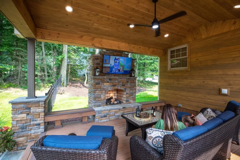 Two people watch TV on a covered patio with a fireplace, wicker furniture, and wooded backyard view.