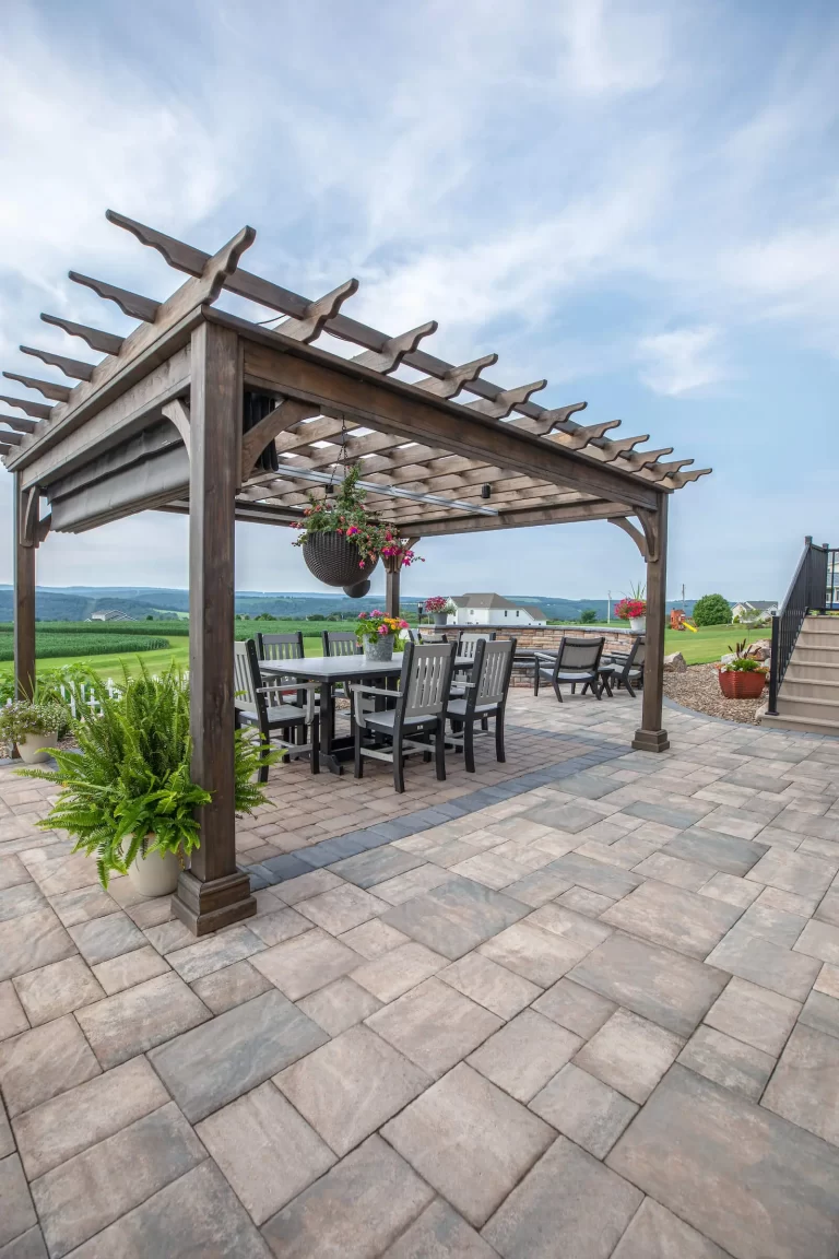 A wooden pergola with a dining set on a paved patio, overlooking green fields and a partly cloudy sky.