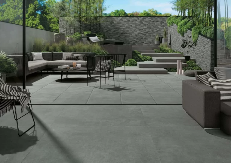 Modern patio with gray tiles, outdoor sofas, chairs, potted plants, and a stone wall with greenery.