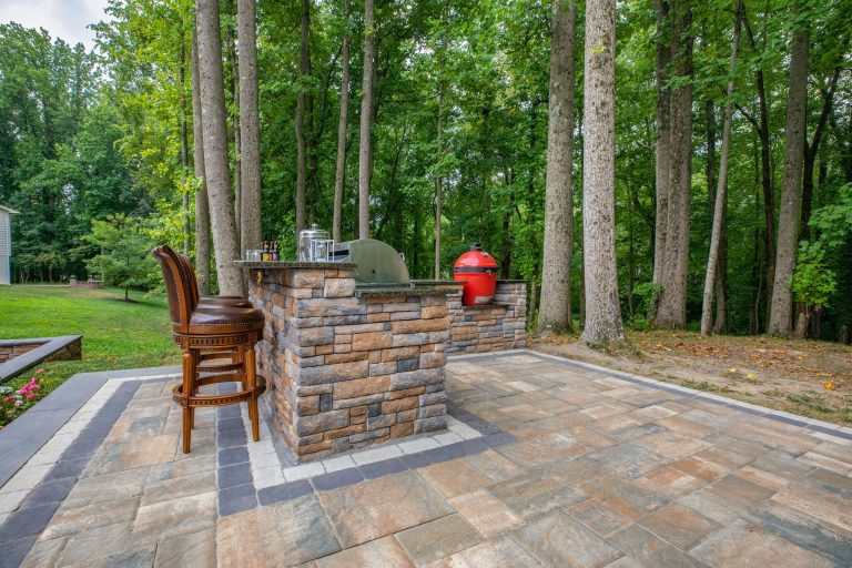 Outdoor kitchen with stone bar, two chairs, and a red grill on a patio surrounded by tall trees.