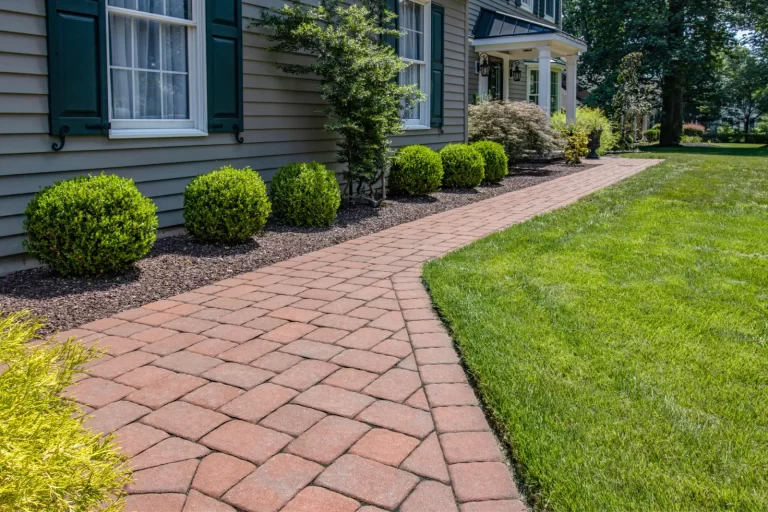 Curved brick walkway beside a house, bordered by green bushes, mulch, and a well-kept lawn.