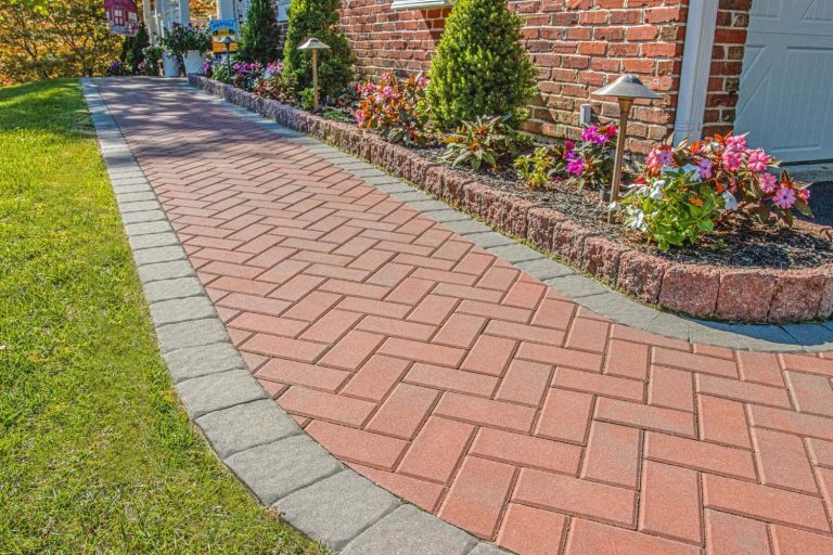 Curved brick walkway bordered by flowers and shrubs beside a brick house on a sunny day.