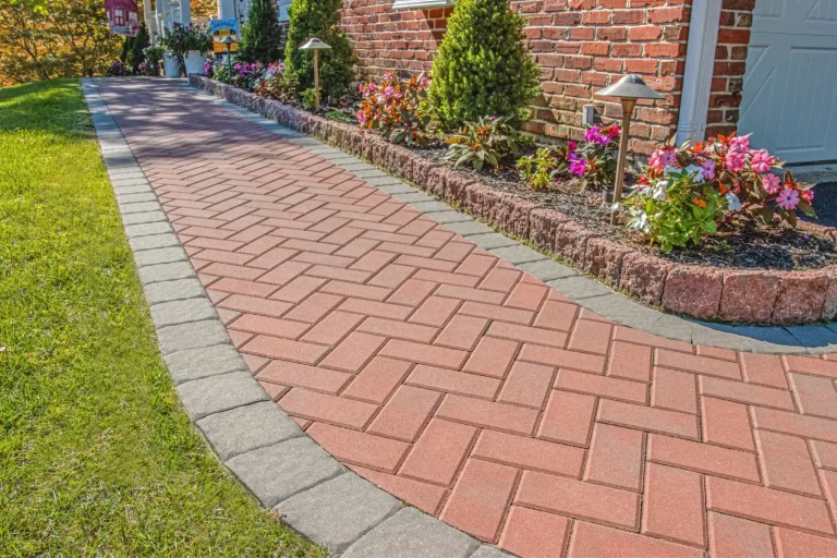 Red brick walkway with gray border, beside garden bed with flowers and small shrubs next to a brick house.
