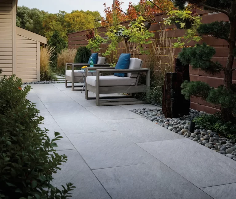 Patio with two cushioned chairs, a small table, and surrounding greenery and rocks along a wooden fence.