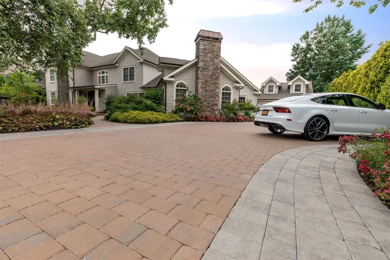 Large house with stone chimney, landscaped yard, and white car parked on a wide paved driveway.