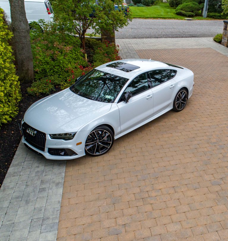 A white Audi sedan is parked on a paved driveway surrounded by greenery and trees.