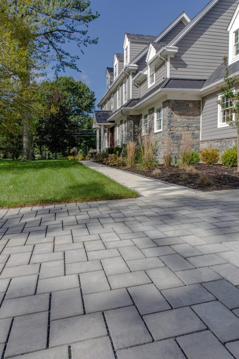 Gray paver walkway leading to a large gray house with stone accents and landscaped yard.