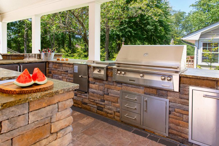 Outdoor kitchen with stainless steel grill, stone counters, and a plate of watermelon on the counter.