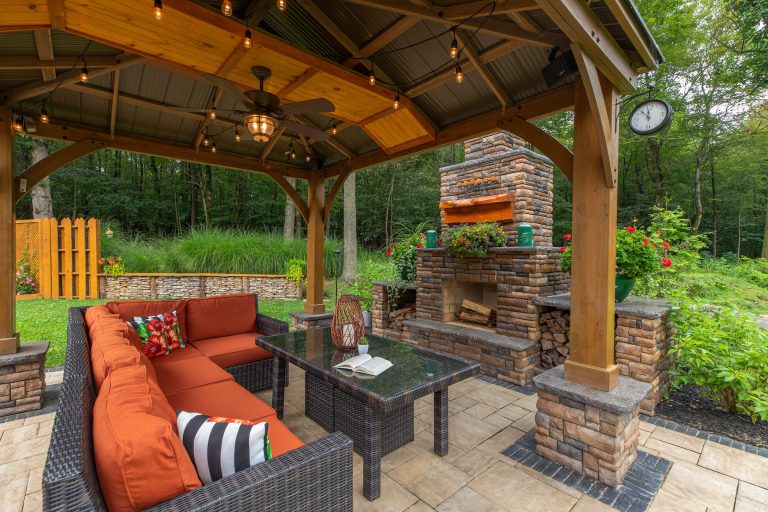 Covered patio with orange cushioned seating, glass table, and stone fireplace in a lush backyard garden.