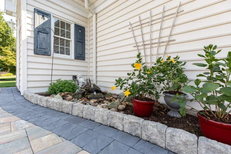 Yellow flowers in pots and a small rock garden beside a white house with a paved stone walkway.