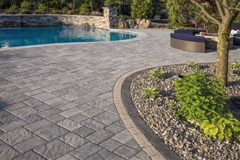 Stone patio with curved garden bed, small plants, and a pool with a stone wall and lounge furniture in the background.