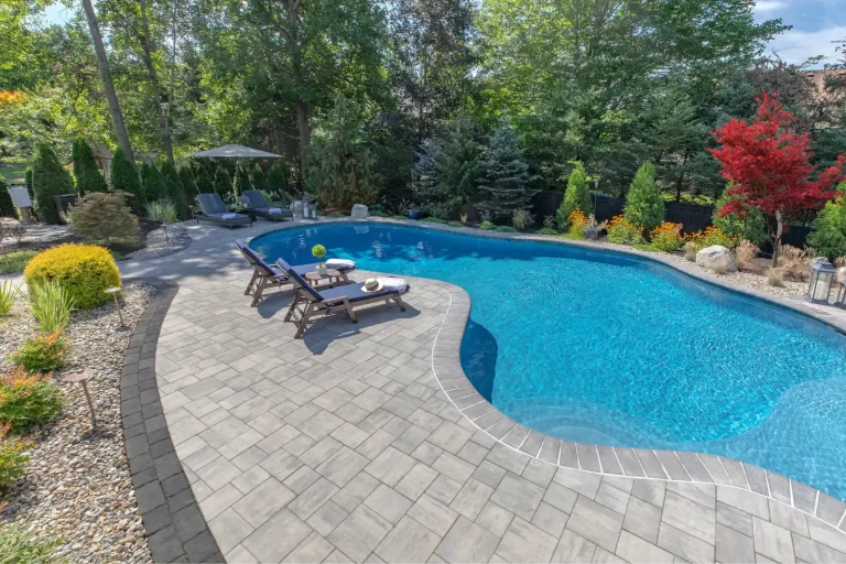 Curved backyard pool with lounge chairs, patio, and lush landscaping surrounded by trees and shrubs.
