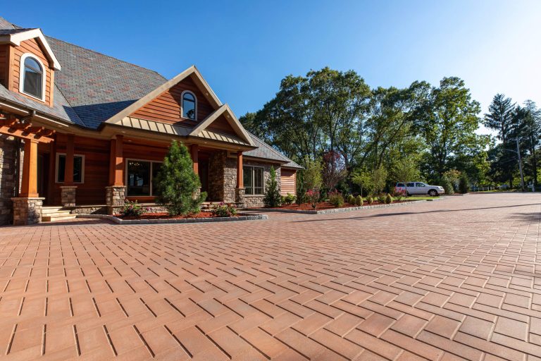 A large brick driveway leads to a wooden house with trees and a parked truck in the background.