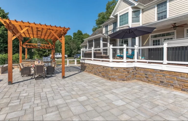 Spacious stone patio with a wooden pergola, outdoor dining set, umbrella, and a house with a porch.
