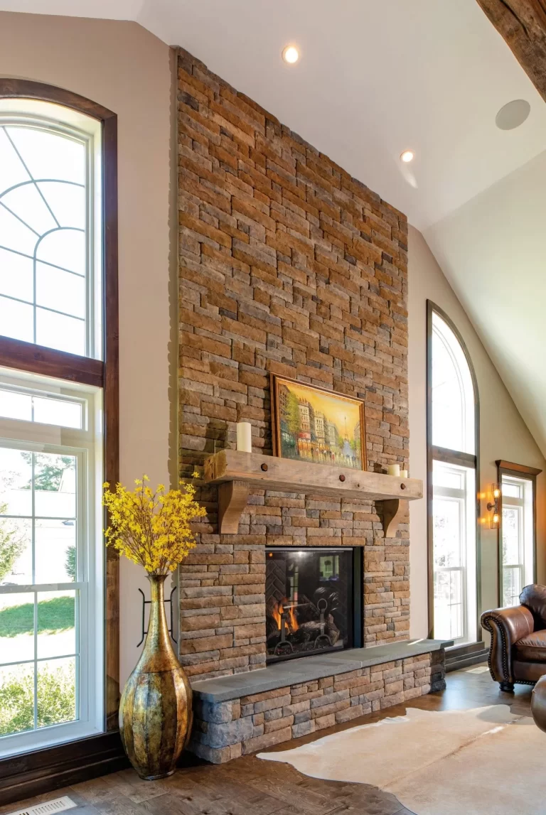 Tall stone fireplace with a wooden mantel, yellow flowers in a vase, and large arched windows on both sides.