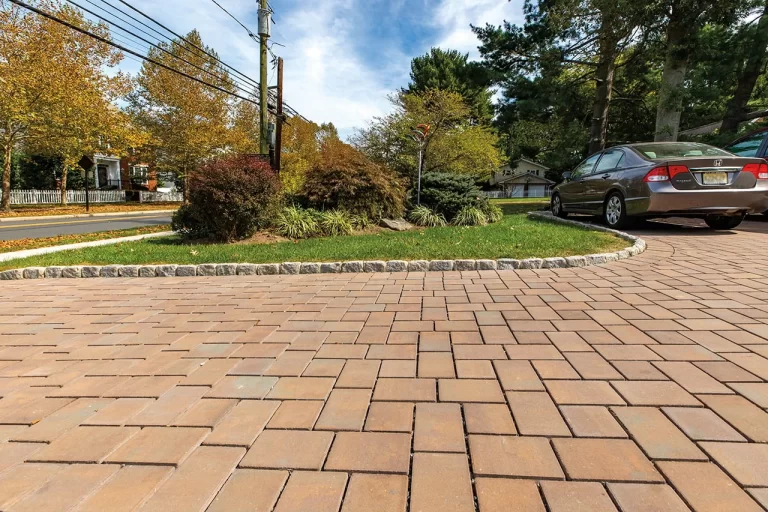 Tan brick paver driveway with a parked car, curb, landscaped greenery, and trees in the background.