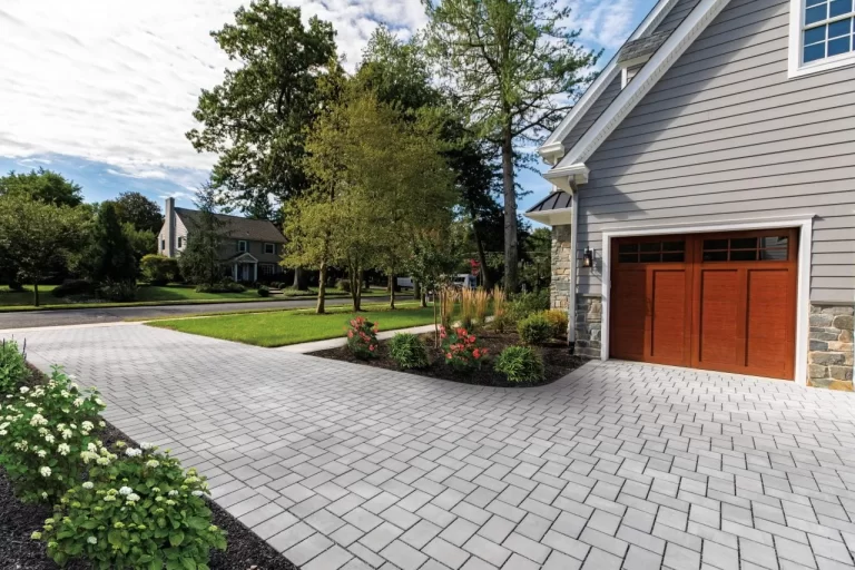 Modern house with a gray brick driveway, wooden garage doors, and landscaped garden with flowers and trees.