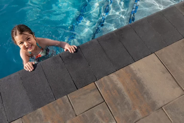 Smiling child holding the edge of a swimming pool, looking up from the water near tiled poolside.