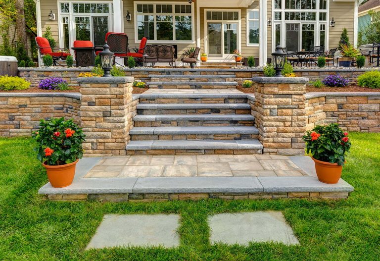 Stone steps lead to a patio with red chairs and potted flowers, surrounded by a green lawn and colorful plants.