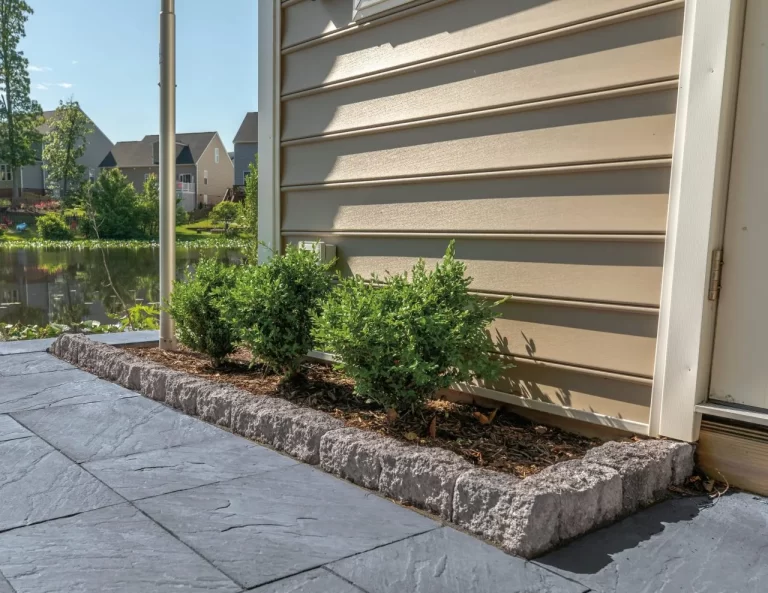Small green bushes planted along a house with beige siding, bordered by stone edging and a stone patio.