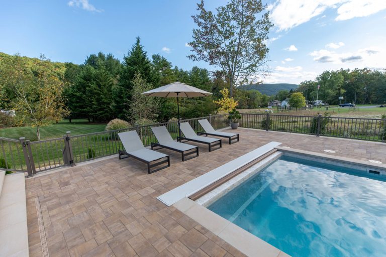 Four lounge chairs and an umbrella are beside a pool on a patio, overlooking a green, tree-filled yard.