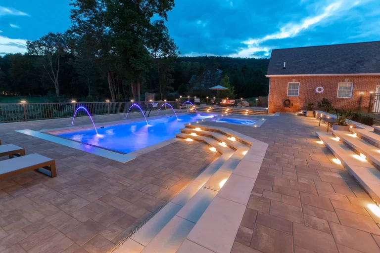 Modern pool with illuminated steps, water jets, and patio seating at dusk near a brick building.