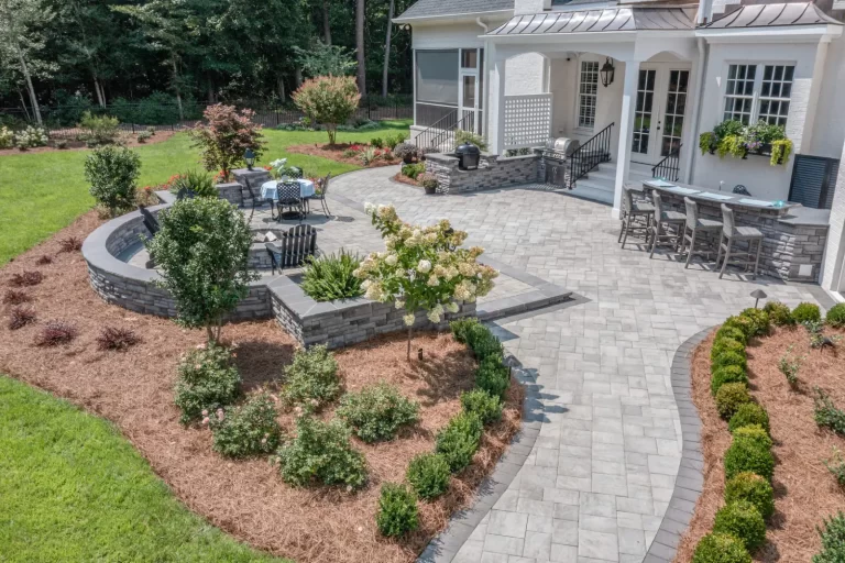 Spacious stone patio with seating, bar area, garden beds, and lush landscaping in a backyard.