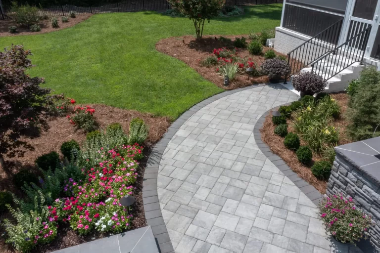 Curved stone walkway bordered by colorful flowers and shrubs leads to a grassy backyard and house steps.