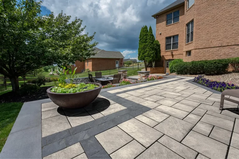 Large paved patio with modern tile design, planter, outdoor seating, and landscaping beside a brick house.