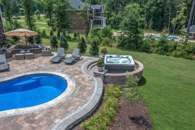 Backyard with pool, hot tub, lounge chairs, outdoor seating, and lawn bordered by trees and houses.
