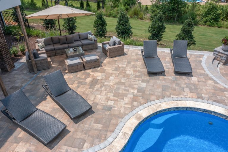 Patio with lounge chairs, sofa, umbrella, and table beside a curved swimming pool and green lawn.