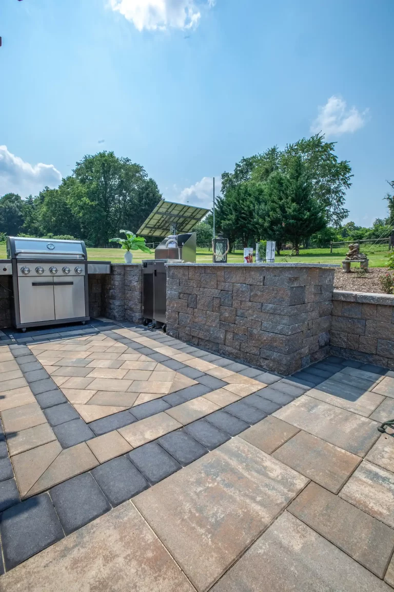 Outdoor kitchen with a stainless steel grill and counter, set on a patterned stone patio with trees in background.