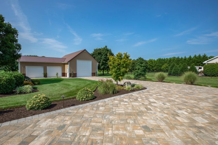 Large driveway with beige pavers, landscaped yard, and a tan garage with white doors under a blue sky.