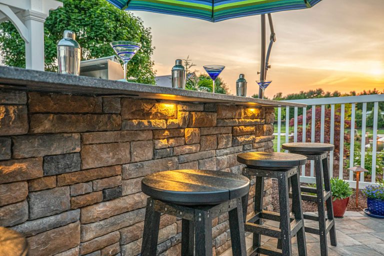 Outdoor stone bar with wooden stools, martini glasses, and drinks, under an umbrella at sunset.