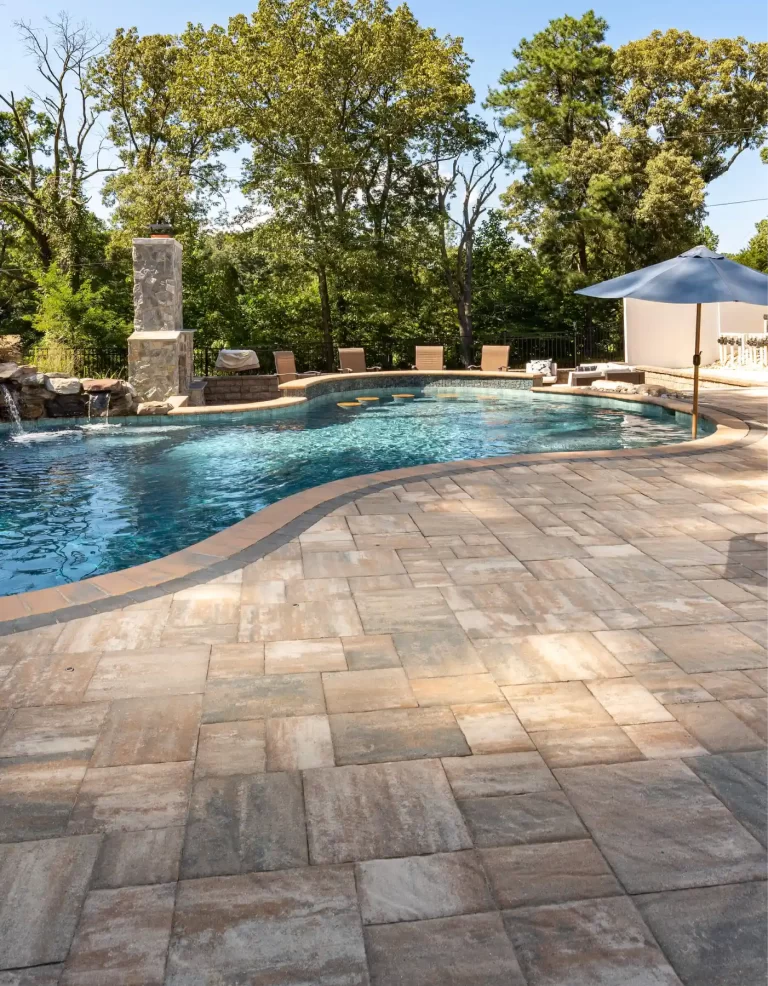 A swimming pool with a stone patio, lounge chairs, an umbrella, and trees in the background.