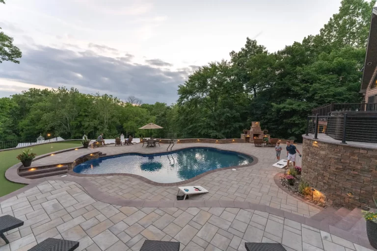 Backyard pool area with a patio, people, cornhole games, outdoor fireplace, and surrounding trees at dusk.