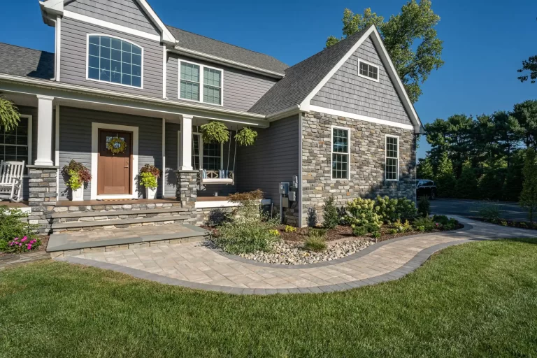 Gray house with stone accents, a landscaped yard, paver walkway, and hanging plants by the front porch.