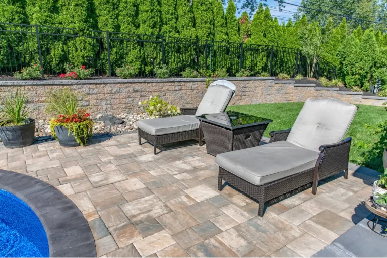 Two wicker lounge chairs with cushions on a stone patio near a pool, surrounded by greenery and potted plants.