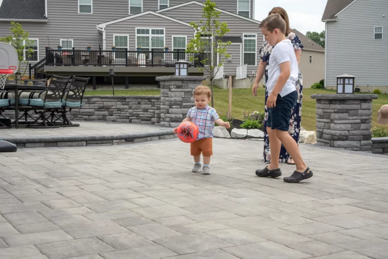 A toddler plays with a red ball outside, joined by a woman and an older boy on a patio.