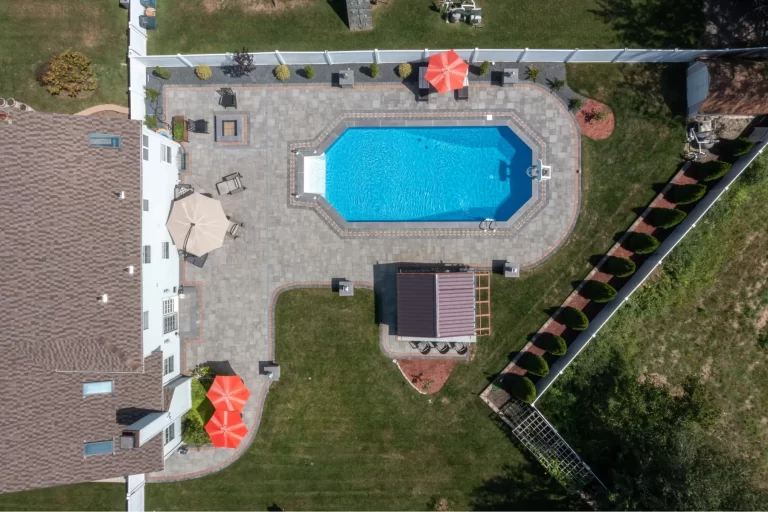 Aerial view of a backyard with a pool, patio, umbrellas, and surrounding green lawn.