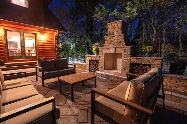 Cozy patio at night with cushioned seating, a stone fireplace, and warm lighting next to a log cabin.