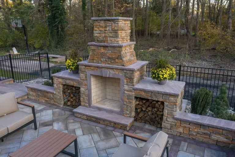 Stone outdoor fireplace with yellow flowers on either side, wood storage, and patio seating in a backyard.