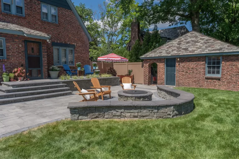 Brick house patio with fire pit, wooden chairs, and lawn; garage and striped umbrella in background.