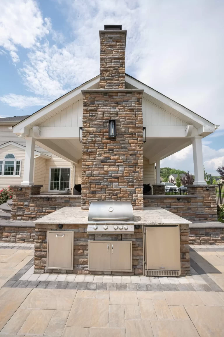 Outdoor stone kitchen with built-in grill and chimney in front of a modern house with covered patio.