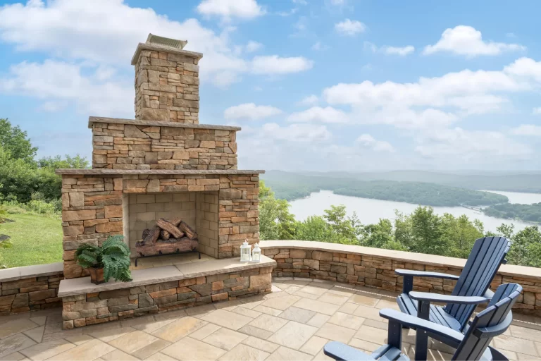 Stone outdoor fireplace on a patio with blue chairs, overlooking a scenic river and lush green hills.