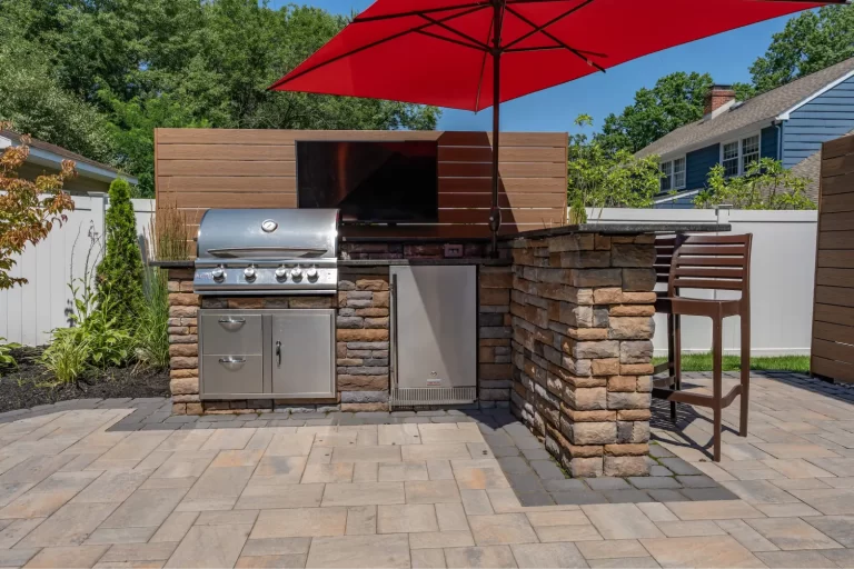 Outdoor kitchen with a grill, mini fridge, TV, and red umbrella on a stone patio.