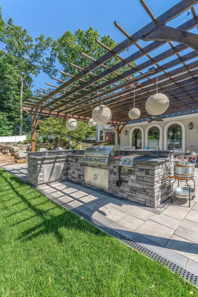 Outdoor kitchen with stone counters, grill, and pergola, hanging lanterns, and green lawn under sunny sky.