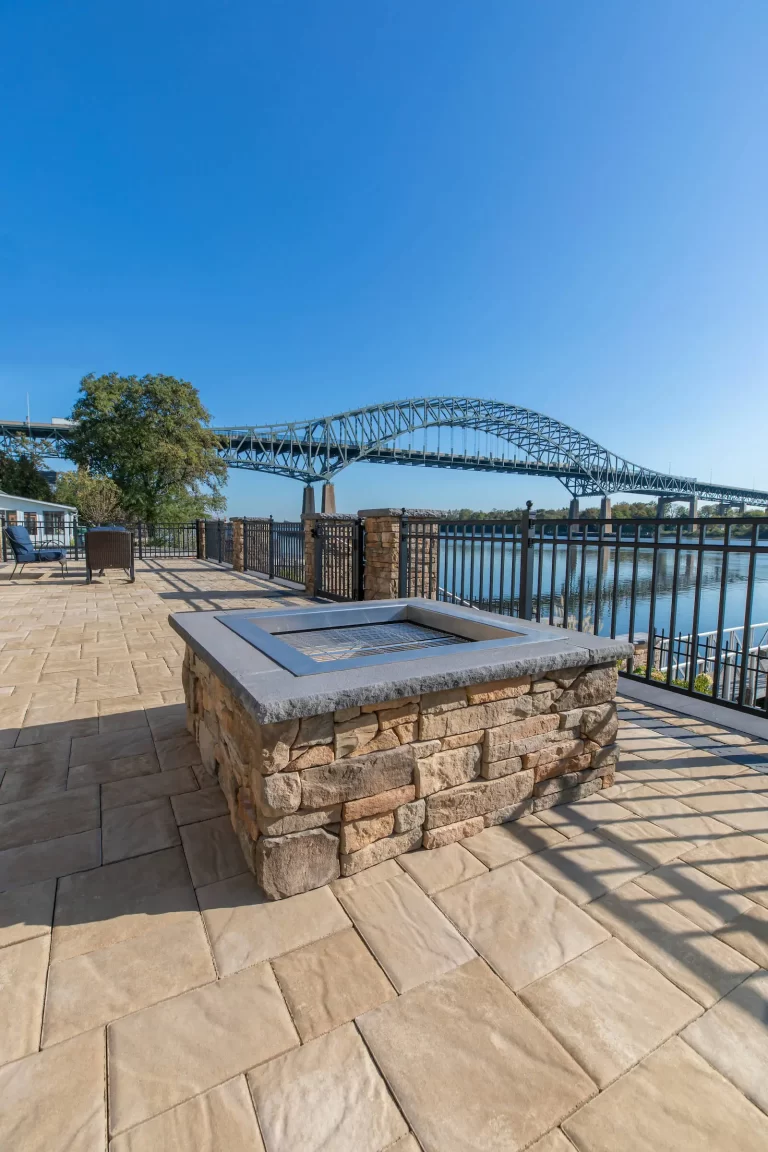 Stone firepit on a patio overlooking a river and a large arched bridge under a clear blue sky.