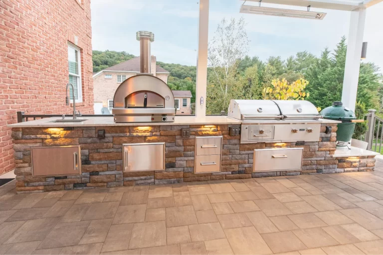 Outdoor kitchen with a brick base, pizza oven, grill, sink, storage drawers, and patio lighting.