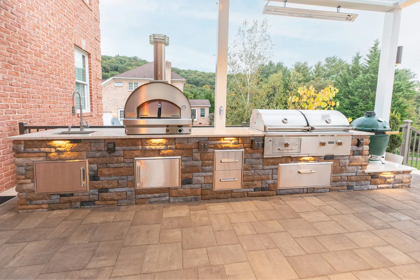 Outdoor kitchen with a brick base, pizza oven, grill, sink, storage drawers, and patio lighting.
